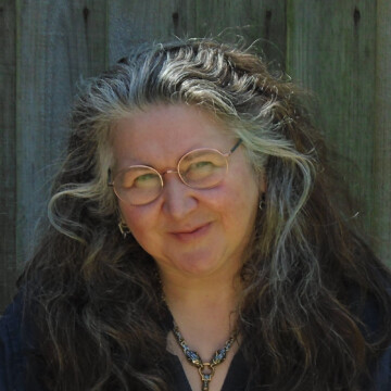 A white woman smiling at the camera. She has long dark hair with grey streaks framing her face and is wearing glasses.