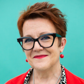 A photo of a white woman smiling. She has short reddish brown hair, black glasses, and red earrings.