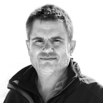 A black and white image of a white man smiling at the camera. He has short hair and is wearing a dark coloured outdoor jacket.