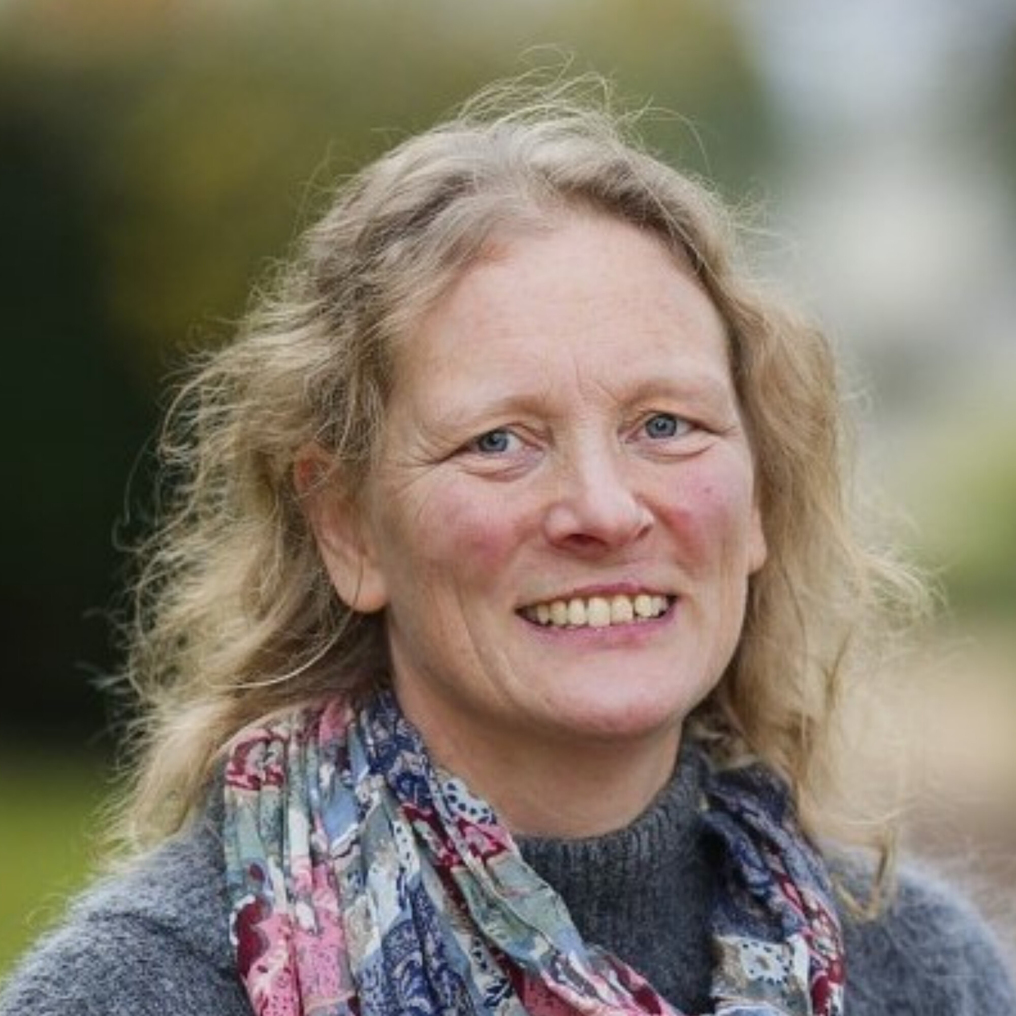 A white woman with blonde wavy hair smiling, wearing a colourful scarf and a blue jumper.