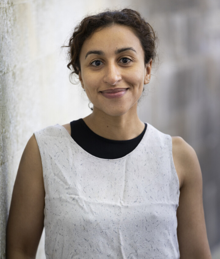 A woman with brown skin and a white linen top is smiling gently at the camera. She has dark hair tied back and is leaning against a wall.