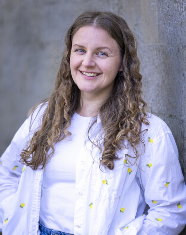 A white woman with light brown curly hair is leaning against a wall and smiling at the camera. She is wearing an oversized white shirt.