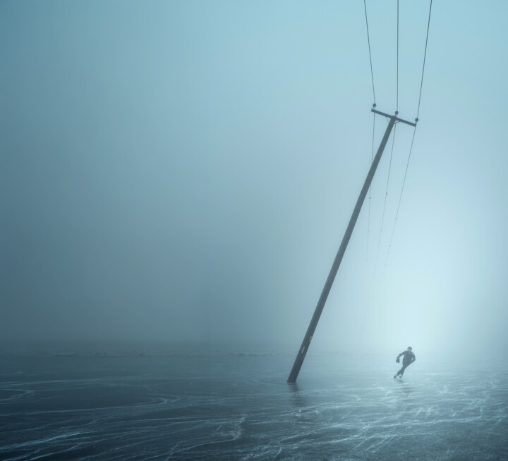 A man ice-skating on the Cambridge fenlands in winter underneath a large, leaning telegraph pole.